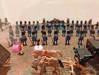 Muñecos de juguete en fila vestidos de soldados azules con sombreros negros, fusiles, caballos y carruajes sobre tierra