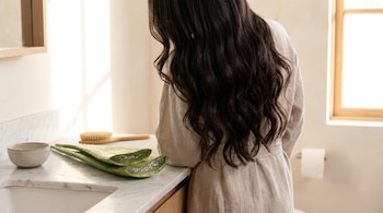 Mujer de espaldas con cabello largo oscuro frente a un mostrador de mármol blanco. Hay hojas de aloe vera cortadas, un cuenco y un cepillo de madera.