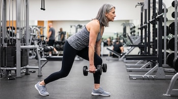 Mujer de cabello gris con ropa deportiva sostiene dos mancuernas mientras realiza un ejercicio físico en un gimnasio.