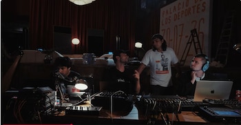 Cuatro hombres. Tres sentados, uno de pie. Consolas de mezcla, dos computadoras portátiles, auriculares, micrófonos, cables, lámparas, cortinas rojas, una pancarta, una escalera