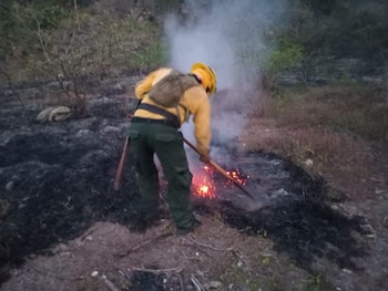 Bombero con casco amarillo, traje amarillo y pantalón verde. Utiliza una herramienta con mango de madera para remover brasas en un suelo quemado con humo
