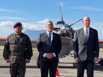 Dos militares con uniforme verde y gorro de cuartel azul están de pie junto a un avión de hélice decorado bajo un cielo nublado