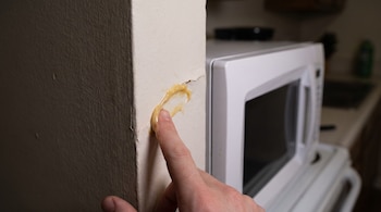 Una mano aplica un gel amarillo sobre la pared blanca junto a un microondas.