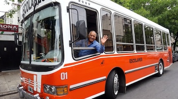 Autobús escolar naranja y blanco con las palabras "ESCOLARES" y el número "01", conducido por un hombre sonriente que saluda desde la ventana.