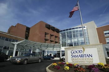 Edificio de hospital con fachada de ladrillo y vidrio, entrada cubierta. Cartel de Good Samaritan Health System. Bandera de Estados Unidos y automóviles