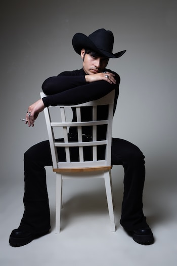 Hombre con sombrero vaquero y ropa negra, incluyendo un cuello alto y botas de plataforma, sentado al revés en una silla blanca, sosteniendo un cigarrillo encendido