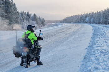 Un hombre con casco y chaqueta verde neón conduce una motocicleta cargada de equipaje por una carretera nevada flanqueada por árboles cubiertos de nieve. El vapor sale del escape