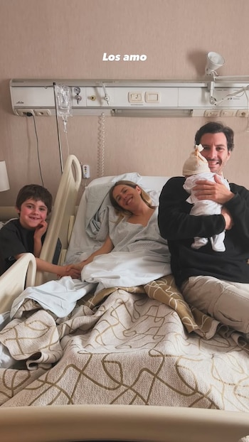 Una mujer en una cama de hospital sonríe, un hombre abraza un bebé y un niño sonríe al lado de la cama. Hay equipos médicos y el texto 'Los amo'