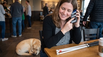 Mujer sonriente usa un teléfono celular en una cafetería junto a su perro guía y bastones blancos plegados sobre la mesa.