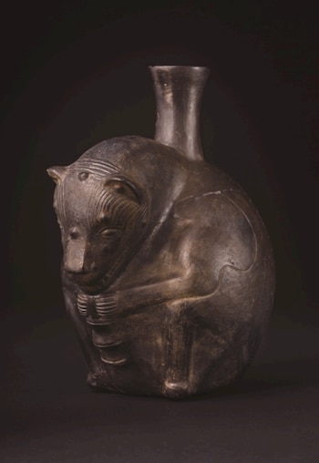Vasija de cerámica oscura representando un perro sin pelo peruano sentado, con detalles grabados en su cabeza y cuerpo, y un cuello estrecho en su lomo. Fondo negro