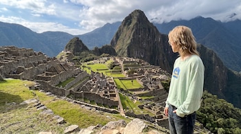 Una persona de perfil con cabello rubio largo y suéter verde observa las ruinas de piedra de Machu Picchu entre montañas.