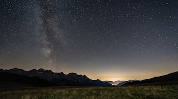 Imagen panorámica de un cielo nocturno oscuro lleno de miles de estrellas, sobre un campo de hierba verde y las siluetas de una cadena montañosa.