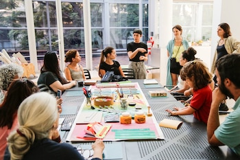 Grupo de personas sentadas y de pie alrededor de una mesa con materiales de manualidades: papeles, tijeras, marcadores, hilos, pegamento y reglas