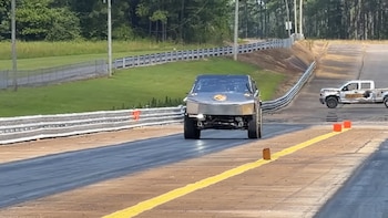 Coche de carreras gris oscuro de diseño angular en pista de aceleración con línea amarilla. Árboles, vallas y una camioneta blanca al fondo