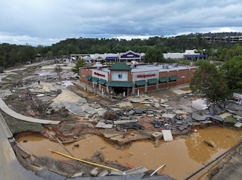 Vista aérea de una zona comercial en Asheville con asfalto roto, grandes socavones llenos de agua fangosa y escombros, y tiendas Walgreens y Lowe's dañadas