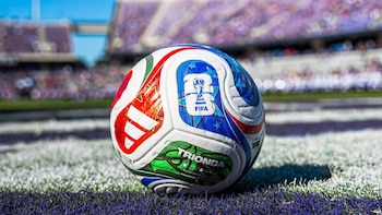 Primer plano de un balón de fútbol blanco con patrones rojos, azules y verdes, y el logo de la FIFA, sobre césped verde y líneas blancas, con un estadio borroso al fondo
