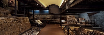 Muros de piedra, escalones antiguos, pasarelas de madera, barandales metálicos, paneles de información y estructuras modernas en un museo
