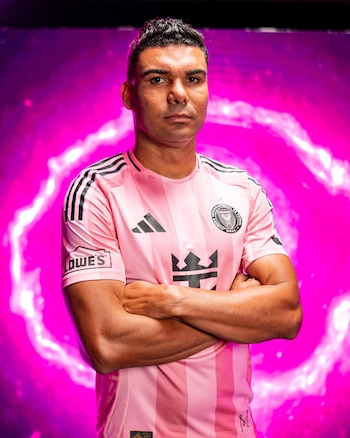 Hombre con cabello oscuro y barba incipiente, viste una camiseta de fútbol rosa con franjas negras y los brazos cruzados, sobre un fondo magenta
