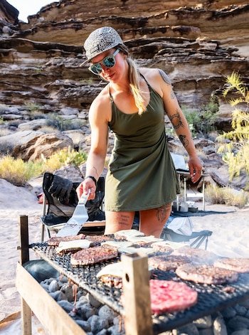Mujer con gorra y gafas de sol asa hamburguesas con queso en una parrilla de carbón; hay sillas plegables, rocas y vegetación