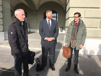 Tres hombres con trajes, abrigos y maletines están de pie sobre una explanada frente a una fachada con arcadas y rejas metálicas