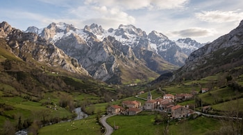 Casas de piedra con tejados rojos en un valle verde junto a un río y altas montañas con nieve en las cumbres.