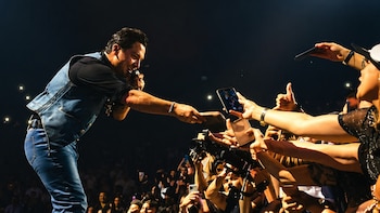 Un cantante masculino en el escenario, con camisa negra y chaleco de mezclilla, se inclina para interactuar con fans que extienden sus manos y teléfonos
