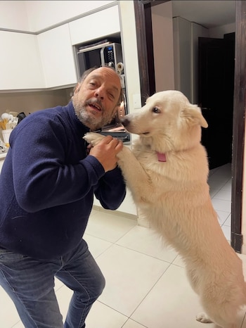 Un hombre con suéter azul sostiene las patas delanteras de un perro blanco parado sobre sus patas traseras en una cocina