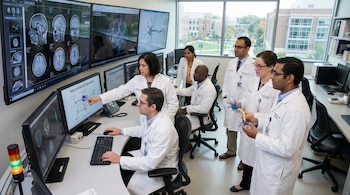 Equipo diverso de científicos en batas blancas en laboratorio, examinando pantallas con imágenes cerebrales, modelos de neuronas y manipulando un modelo físico de neurona.