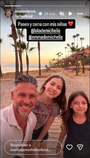 Martín Demichelis sonriendo junto a dos niñas. De fondo, una pasarela de madera, arena, mar, palmeras y edificios costeros bajo un cielo anaranjado