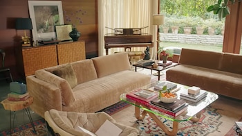 Salón con sofás beige, mesa de centro de cristal con libros y fruta, piano, tocadiscos y ventanales que dan a un jardín exterior