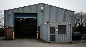 Un galpón industrial gris con portón azul abierto, un contenedor de seguridad metálico con puerta y ventanas de reja, y varias cámaras de vigilancia.
