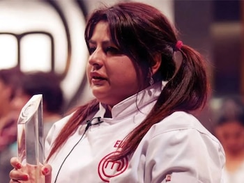 Una mujer con chaqueta de chef blanca y el logo de MasterChef, cabello castaño rojizo en dos coletas, sostiene un trofeo de cristal