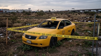 Un taxi amarillo con el parabrisas roto rodeado por cintas amarillas de la policía en un terreno baldío.