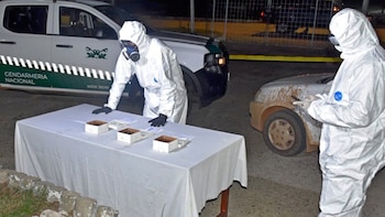 Duas pessoas com roupas e luvas de proteção brancas, uma delas com máscara de gás, examinam as bolhas na mesa. Atrás, um carro da polícia e um carro