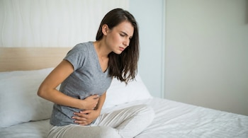 Mujer joven sentada en una cama con sábanas blancas, vestida con camiseta gris y pantalones claros, agarrándose el abdomen con ambas manos.