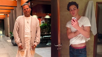Dos hombres. Uno con cárdigan, gafas y perro en pasillo. Otro con camiseta y vaqueros, toma selfie con teléfono en baño con toalla.