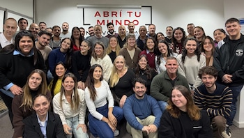Un grupo grande de hombres y mujeres de diversas edades sonríen frente a una pared blanca con un cartel que dice 'ABRÍ TU MUNDO' en letras rojas