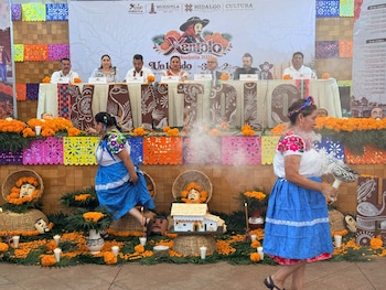 Un panel de autoridades tras una mesa decorada con la palabra Xantolo, flores de cempasúchil, artesanías y dos mujeres con trajes regionales
