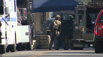 Agente del FBI en equipo táctico junto a vehículos de emergencia, incluyendo un blindado y una SUV roja, en calle durante operativo policial