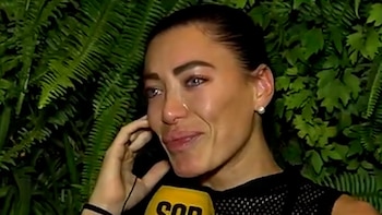Primer plano de Pamela Pombo, con lágrimas en los ojos, sosteniendo un micrófono amarillo mientras mira ligeramente hacia la derecha. Fondo verde de follaje