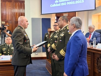 Primer plano de un oficial militar calvo en uniforme verde oliva sosteniendo un documento o placa, frente a un hombre de traje azul y otros oficiales