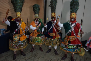 Cuatro personas con atuendos tradicionales coloridos, que incluyen máscaras, tocados de plumas de pavo real, túnicas bordadas y objetos de madera