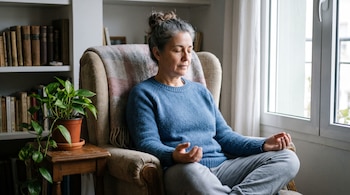 Mujer con los ojos cerrados sentada con las piernas cruzadas en un sillón junto a una ventana, una planta pequeña y estantes con libros.