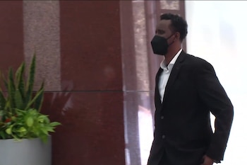 Un hombre con traje oscuro y mascarilla negra camina en un pasillo junto a una pared de mármol y una planta decorativa