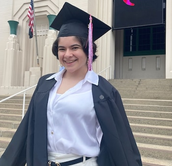 Joven con toga y birrete de graduación negro con borla rosa, sonríe ante un edificio con una bandera y escaleras. Es Gabriella Pizzolo