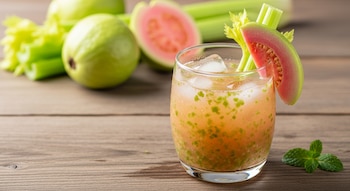 Un vaso de agua de guayaba y apio con hielo, decorado con una rodaja de guayaba rosada y un tallo de apio, sobre madera. Guayabas, apio y menta fresca se ven al fondo.