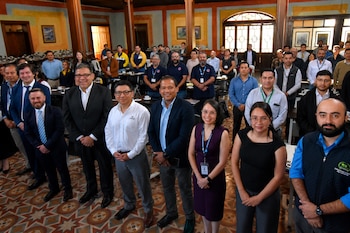 Un grupo de personas vestidas de forma formal y semiformal posa de pie en un salón con columnas de madera y suelo decorado.