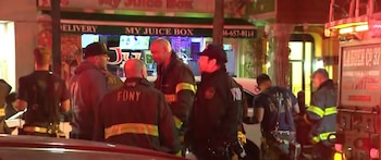 Varios bomberos y agentes de policía se reúnen en una calle por la noche, con un camión de bomberos y un negocio 'My Juice Box' iluminado al fondo