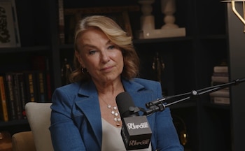 Primer plano de una mujer de mediana edad con cabello rubio, blazer azul y collar de perlas, sentada frente a un micrófono en un estudio oscuro con estanterías