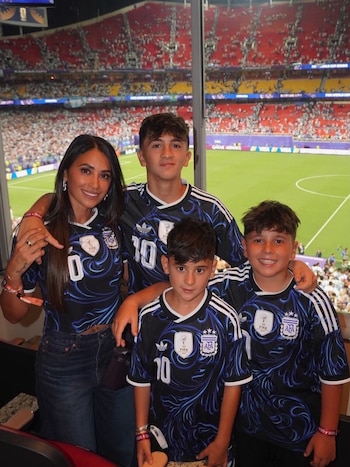 Antonela Roccuzzo con tres niños sonríen y posan en un palco de estadio. Visten camisetas de la selección argentina. Al fondo, la cancha y tribunas llenas